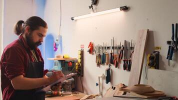 Craftsperson drawing blueprints on notepad in studio with BIPOC coworker in background solving tasks. Manufacturer looking at technical schematics to execute woodworking projects, camera B photo