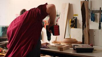 Woodworker in assembly shop using power drill to create holes for dowels in wooden board. Carpenter sinking screws into wooden surfaces with electric tool, doing precise drilling for seamless joinery, camera A photo