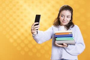 Smiling woman holding pile of books, taking selfies with smartphone, conveying joy of reading concept, studio background. Cheerful person with novels taking pictures with phone photo