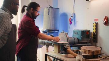 Manufacturer teaching new employee how to shape wooden bowl on disc sander in assembling shop. Apprentice learning from woodworking specialist how to use grinding machine rotating disc, camera B photo