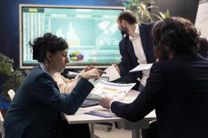Business team evaluate financial reports and success metrics on papers, using sales infographics for annual data reports. General director and staff members studying market demands and needs. photo