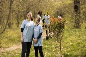 retrato de mamá y su hija trabajar como voluntario a planta arboles en el bosque área, Ayudar con repoblación forestal y conservación de el natural ecosistema. sentido de devoción y responsabilidad. foto