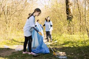 Little child activist collecting rubbish in blue garbage disposal bags, cleaning the natural forest environment and recycling trash. Ecologic volunteer grabbing plastic waste and litter. photo