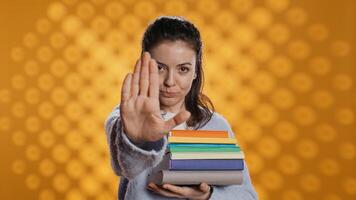 Portrait of stern woman holding stack of books doing stop sign gesturing, studio background. Student with pile of textbooks in arms used for academic learning doing halt hand gesture, camera B photo