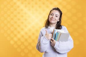 Portrait of happy woman carrying stack of textbooks, gathering information for school exam, studio background, showing thumbs up. Jolly person holding pile of books, doing positive hand gesturing photo