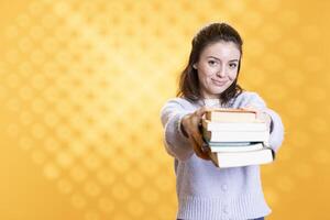Portrait of friendly woman offering stack of textbooks useful for school exam, isolated over studio background. Merry person giving pile of books, recommending them for university assessment photo