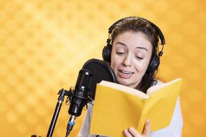 Cheerful woman with headphones on narrating book word for word to produce audiobook. Jolly narrator using storytelling skills and microphone to produce digital recording of novel, studio background photo