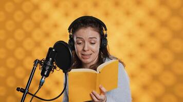 Happy narrator wearing headphones reading aloud from book into mic against yellow background. Joyous lady recording audiobook, creating engaging media content for listeners, camera A photo