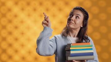 Portrait of smiling woman pointing towards empty copy spaces while holding pile of books. Radiant person with stack of novels in arms promoting reading, showing messages, studio background, camera A photo