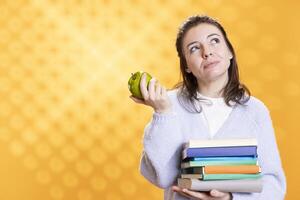 Portrait of relaxed woman daydreaming while holding pile of books, eating apple. Person with stack of novels in arms caught in imagination reverie, enjoying healthy fruit, studio background photo