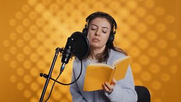 Narrator wearing headset reading aloud from book into mic against yellow background. Upbeat professional voice actor recording audiobook, creating engaging media content for listeners, camera B photo