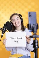 Smiling woman holding placard with world book day message written on it, filming with phone, studio background. Happy content creator recording promoting importance of reading photo