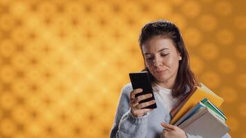Smiling woman reading messages on smartphone while holding stack of books, isolated over studio background. Cheerful person with pile of academic textbooks in arms texting friends on phone, camera A photo
