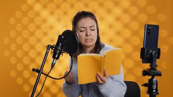 Online star films vlog at work using smartphone on tripod, passionately reading book to record audiobook. Narrator doing content creation with phone, producing digital recording of novel, camera B photo