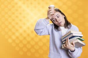 Woman feeling migraine from stress, carrying books for school exam and coffee cup to wake up, studio background. Exhausted student suffering headache from too much homework, drinking caffeine beverage photo