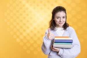 Portrait of happy woman with stack of books in hands showing thumbs up, isolated over studio background. Joyous bookworm holding pile of novels, feeling upbeat, doing positive hand gesturing photo