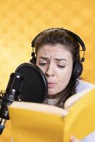 Woman frowns while doing voiceover reading of book to produce audiobook using melodramatic acting. Narrator glowers, using storytelling skills to portray character, recording novel, studio background photo