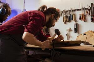 Experienced woodworker shaping raw timber using chisel and hammer in carpentry shop, wearing safety glasses. Woodworking expert shaping wooden objects, making wood sculptures using protective gear photo