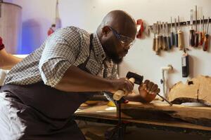 BIPOC man in carpentry shop wearing protection equipment while carving wood to prevent workplace accidents. Cabinetmaker equipped with safety goggles while using chisel and hammer to avoid injury photo