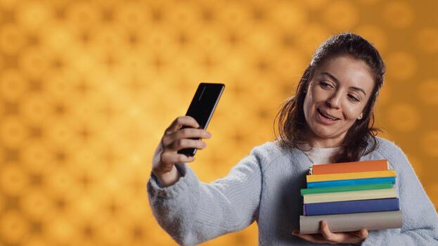 Reading enthusiast woman filming social media videos with smartphone holding stack of books, studio background. Cheerful nerd with pile of textbooks in arms recording with selfie camera, camera A photo
