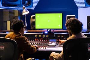 Team of musicians editing music with greenscreen on computer in professional recording studio, using audio mixing console with knobs and faders in control room. Artist and producer compose tracks. photo