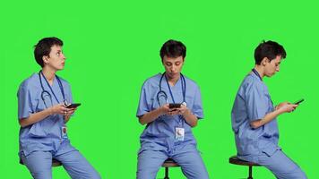 Nurse browsing online websites on smartphone app in studio, sitting on a chair against greenscreen backdrop. Medical specialist in scrubs using mobile phone and social media texting. Camera B. photo
