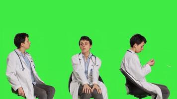 Physician in white coat waiting for patients at consultations, feeling impatient sitting on a chair against greenscreen backdrop. Medic practitioner with stethoscope waits for people. Camera B. photo