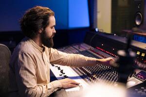 Music producer editing and mixing tunes in control room, working on a new song in professional recording studio. Audio engineer using control panel board with buttons and motorized faders. photo