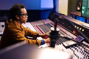 African american audio engineer mixing and mastering music with professional audio console in control room. Technician expert uses soundboard pads on mixer station, post production. photo