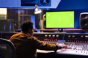 Music engineer working on mockup pc and mixing console with various knobs and faders in control room, editing tunes in post production. Young skilled producer creating sounds, technical equipment. photo