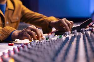 African american sound engineer twisting knobs to edit music in studio, operating control panel board desk with motorized faders and buttons. Skilled producer creating records with gear. Close up. photo