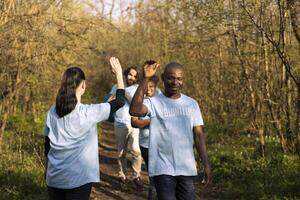 Cheerful satisfied activists sharing high five with each other in the woods, finishing to tidy up the forest ecosystem against pollution. Pleased volunteers showing praise after a job well done. photo