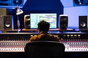 African american record producer editing and mixing tracks on audio console, using digital software on pc to add sound effects in post production. Technician expert works in professional studio. photo