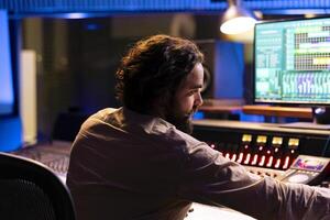 Sound technician producing new music with mixing console, editing tunes with digital software on computer. Skilled music producer processing sounds in control room, uses panel board. photo