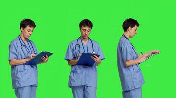 Confident hospital nurse taking notes for the next medic appointment, standing over greenscreen backdrop. Medical assistant with uniform writing patient information on clipboard papers. Camera B. photo