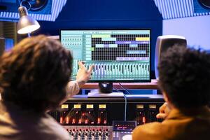 Audio producer and musician working on music for new album, composing and editing songs with mixing console and processing software on pc. Artist working with expert for tunes in studio. photo