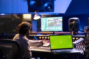 Sound engineer working on control desk with greenscreen layout in professional studio, using sliders and knobs to edit music for album. Audio technician operates soundboard panel for tracks. photo