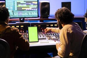 Singer and audio expert working with isolated screen on gadget, mixing and mastering tunes with mixing console and digital software interface. Artists team editing music in professional studio. photo