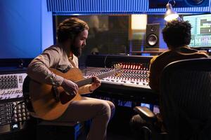 Singer creating a new hit song in studio with guitar running, working with audio expert to produce music. Songwriter singing a record on electro acoustic instrument in control room with soundboard. photo
