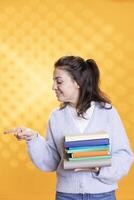 Portrait of upbeat person with stack of books in hands pointing towards empty copy space, isolated over studio background. Cheerful bookworm holding pile of novels, pointing finger off camera photo