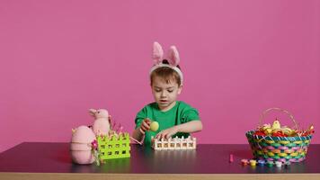 Happy pleased child decorating easter arrangements with handcrafted items, arranging painted colorful eggs in a basket. Joyful little boy celebrating spring festivity, pink backdrop. Camera B. photo