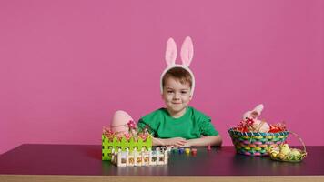 Sweet little child putting fluffy bunny ears on his head and smiling on camera, happy innocent boy sitting at a table with art and craft supplies to decorate easter eggs. Spring holiday. Camera B. photo