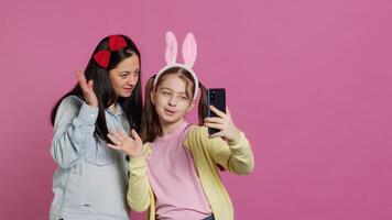 Little schoolgirl and her mom talking on videocall meeting in studio, waving at webcam and chatting with their family. Cute preteen and mother using smartphone for a teleconference chat. Camera A. photo