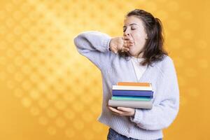 Woman yawning while holding pile of books, tired after spending all day on reading hobby. Bookworm with stack of novels in arms feeling exhausted from lack of sleep, studio background photo