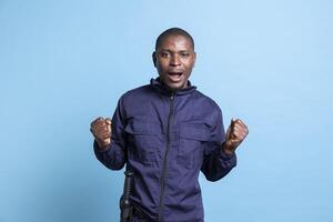 African american agent with uniform acting shocked in studio, having a surprised reaction after seeing something shocking. Male security guard acting amazed against blue background. photo