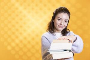 Woman holding stack of books, pointing it towards camera, isolated over yellow studio background. Portrait of smiling bookworm lady presenting stack of novels, urging people to read photo