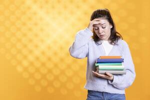 Portrait of upset woman holding pile of books, doing hand on temples gesture, conveying hate of reading concept, studio background. Irked lady with stack of novels showing distaste for literature photo