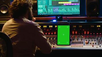 Music producer recording, mixing and reproducing sound on audio software, using stereo console and switchers to increase volume level and enhance tunes. Greenscreen display on tablet. Camera A. photo