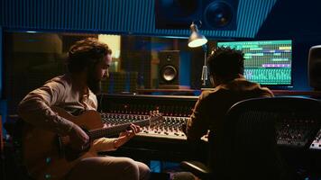 Composer musician recording a new song on his electro acoustic guitar in professional studio, using mixing console and amplifier in control room. Young artist composing new tracks. Camera B. photo