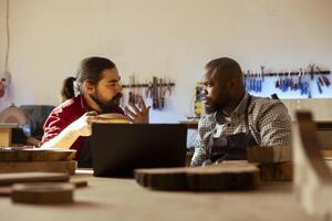 Woodworker looking over blueprints on laptop, brainstorming with african american apprentice. Manufacturer and coworker discussing furniture assembly schematics on notebook in studio photo